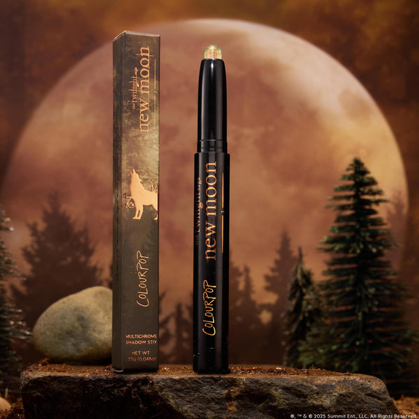 Wolf Girl ColourPop Shadow Stix stands uncapped beside its wolf-graphic box, showcasing a shimmering multichrome eyeshadow tip on a rocky surface with pine trees and a full moon backdrop.