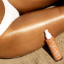 ColourPop SOL Shimmering Dry Oil in Warm Gold - pump bottle on beach sand - highlights a sun-kissed leg in a white swimsuit, with golden shimmer streaking across the skin to showcase the luminous, fast-drying body oil finish.