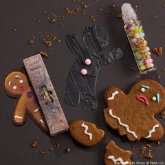 ColourPop Candy Lip Oil in Gumdrop Buttons flavor rests beside its Shrek box, the rollerball tube featuring Gingy art and pastel gumdrop beads, styled on a dark surface with broken gingerbread cookies and crumbs.