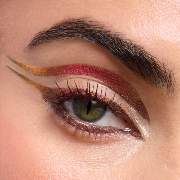 Eye showcasing Dragon’s Keep Crème Gel Liner by ColourPop Crème Gel Liner in a bold double-wing - metallic red-to-copper arc above the crease, olive-gold swoop extending outward, and shimmering rose along the lashline - on fair skin with a dark brow.