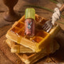 ColourPop Ultra Glossy Lip in Noble Steed rests on syrup-drizzled waffles, the compact gloss bottle featuring a lime-green cap and Donkey graphic label, styled on a rustic wooden board with a fork for a playful breakfast-themed product shot.