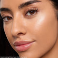 Model wearing Ohana ColourPop Super Shock Highlighter catching light on the cheekbone, delivering a pearly champagne glow with a smooth, dewy finish; close-up beauty shot shows natural skin texture, soft lighting, glossy lips, and defined brows.