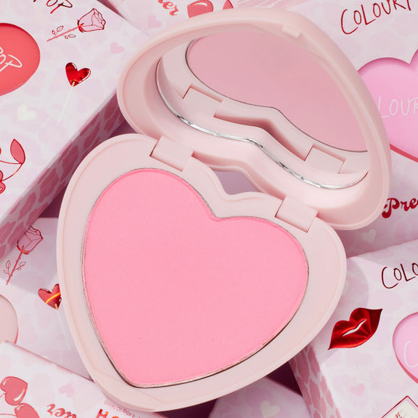 Sweetheart heart-shaped compact opens to reveal a soft baby pink pan and curved mirror, ColourPop Pressed Powder Blush, resting on Valentine-themed packaging with hearts, roses, and lip prints.