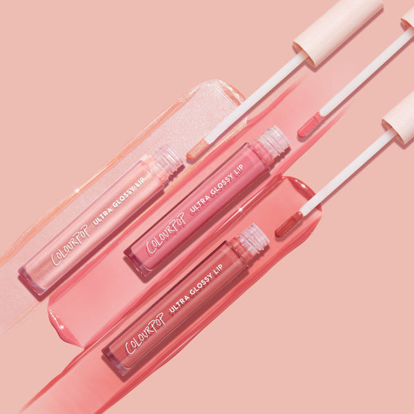 Three ColourPop Ultra Glossy Lip tubes in Tickled Pink glide swatches with doe-foot applicators, showcasing a sheer shimmer, rosy pink, and warm nude finish on a blush pink backdrop; hydrating high-shine Ultra Glossy Lip trio.