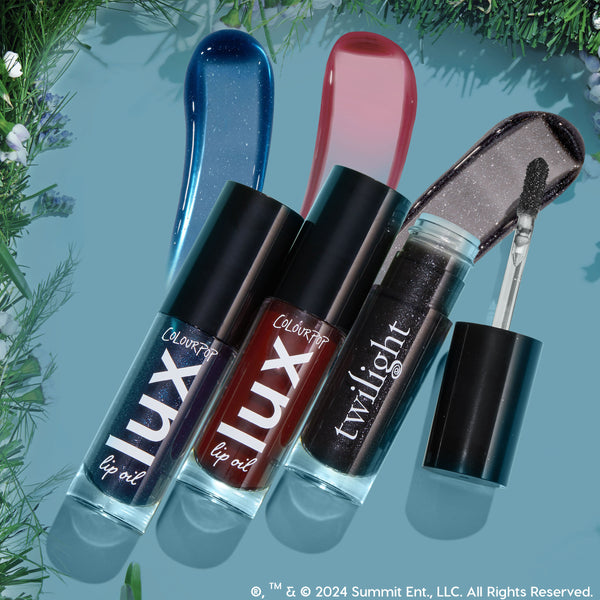 Vampy Lips ColourPop Lux Lip Oil trio in blue shimmer, deep red, and black sparkle shades, shown with glossy swatches and an open doe-foot applicator, bottles angled on a teal backdrop bordered by greenery.