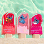 Three ColourPop Ultra Glossy Lip Keepin’ It Sweet tubes stand upright in beach sand, popsicle-shaped casings with drippy gloss and Disney Lilo & Stitch graphics in red, blush pink, and hot pink, set against turquoise pool water.