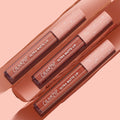 Three ColourPop Ultra Matte Lip tubes in the Warmed Up bundle rest diagonally atop wide nude‑brown swatches, showcasing a full‑coverage ultra matte finish with clear barrels and blush pink star caps on a peach backdrop.