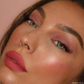 Rosy cheeks wearing Current Crush from the ColourPop Heart Powder Blush Set, blush swept on the high points with a soft satin glow, paired with mauve eyeshadow and coral-pink lips in a close-up against a peach backdrop.