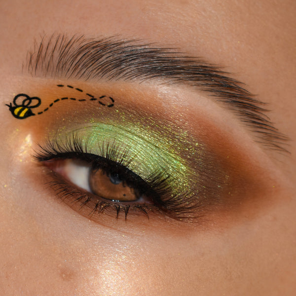 Shimmering green-gold eye makeup blends metallic lime on the lid into warm honey-brown matte, accented by a tiny bee graphic near the brow, on a close-up eye with long lashes, showcasing the Sweet As Can Bee Shadow Palette, a ColourPop Shadow Palette.