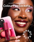 Baby Fox ColourPop Lip Stain coating a model’s smiling lips while a magenta tube is held beside a pink phone with a heart PopSocket, highlighting the ColourPop x PopSockets collab in a close-up with floral nails and pink eye makeup.