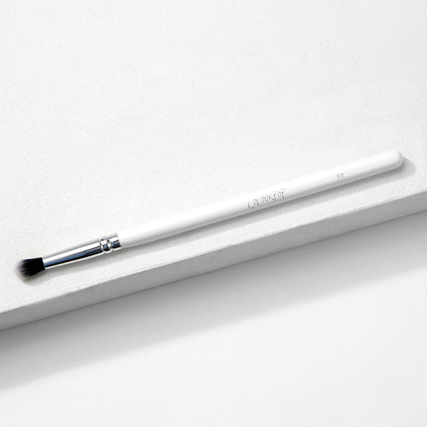 ColourPop Small Tapered Brush resting diagonally on a white textured platform, featuring a slim white handle, chrome ferrule, and small tapered black-and-white bristles for precise eyeshadow blending and detail work.