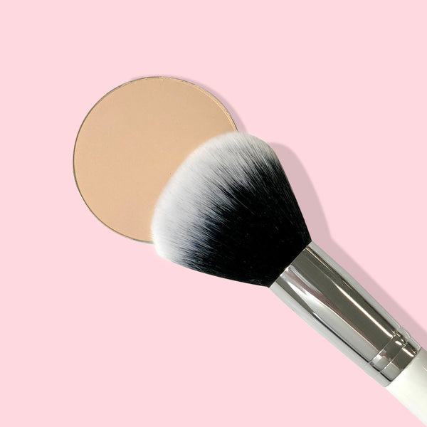 ColourPop Large Powder Brush buffing a beige pressed powder pan, soft black-to-white bristles with shiny silver ferrule and white handle, makeup brush for allover face setting on a pastel pink background.