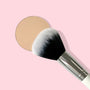 ColourPop Large Powder Brush buffing a beige pressed powder pan, soft black-to-white bristles with shiny silver ferrule and white handle, makeup brush for allover face setting on a pastel pink background.