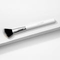 ColourPop Angled Face Brush makeup brush resting diagonally on a white textured platform, featuring a white handle with ColourPop logo, shiny silver ferrule, and soft black-to-white angled bristles for precise blush, bronzer, and contour.