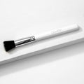 ColourPop stippling makeup brush resting diagonally on a white textured block, featuring duo-fiber black-and-white bristles, a shiny silver ferrule, and a white handle with the ColourPop logo.