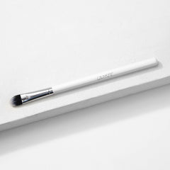 ColourPop Medium Shader Brush with flat oval, dual-toned black-to-white synthetic bristles and a silver ferrule on a white handle, resting on a white textured platform in studio lighting; makeup brush for precise eyeshadow packing and blending.