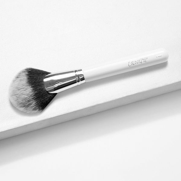 ColourPop Full Fan Face Brush with wide fanned bristles sweeping powder or highlighter, resting at an angle on a white textured surface, featuring a glossy white handle, silver ferrule, and black-to-white bristle gradient.