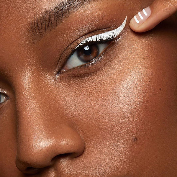 White winged eyeliner defining the upper lash line, applied with ColourPop BFF Liquid Liner in Graceland; close-up of one eye with a fingertip pulling the outer corner, showing a crisp, matte line on brown skin for a bold graphic look.