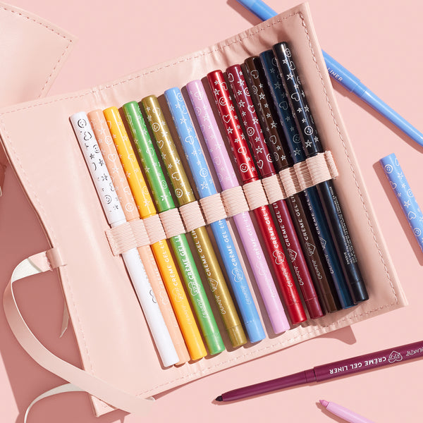ColourPop Crème Gel Liner It’s a Vibe set shows 15 multicolor liners neatly held by elastic in a blush pink roll-up pouch, playful icon prints on barrels, with a few loose pencils around on a pink surface.