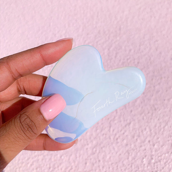 Opalite Crystal Gua Sha facial massage tool held in a hand, showing its iridescent blue‑white stone and curved edges, branded Fourth Ray Beauty, against a pink textured background; part of ColourPop Skincare Tools And Accessories.