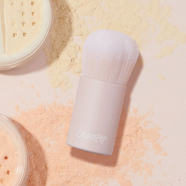 Mini Kabuki Face Brush buffing loose setting powder, a short-handled ColourPop Makeup Brush with dense white bristles and a blush-pink barrel, resting amid peach and yellow powders beside open sifter jars, ideal for travel and seamless blending.