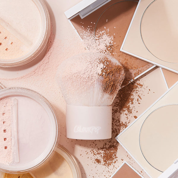 ColourPop Kabuki Face & Body Brush buffs loose and pressed powder, dense rounded bristles coated with pink and tan pigments, ColourPop Makeup Brush resting on a vanity amid open compacts and jars in light-to-tan shades.