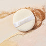 Powder Puff pressing into loose setting powder, a soft round ivory pad with COLOURPOP logo strap resting on swirls of beige, tan, and pink powders - an essential from ColourPop Makeup Tools & Accessories for smooth blending and shine control.