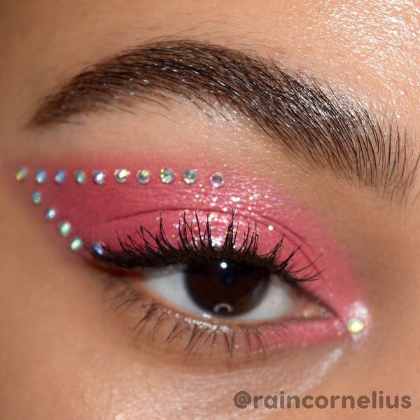 Individual ColourPop Crystal Face Jewels accent a brown eye, forming a dotted wing above shimmery pink eyeshadow with an inner-corner gem; iridescent adhesive rhinestones line the upper lid and outer edge, showcasing placement and sparkle.