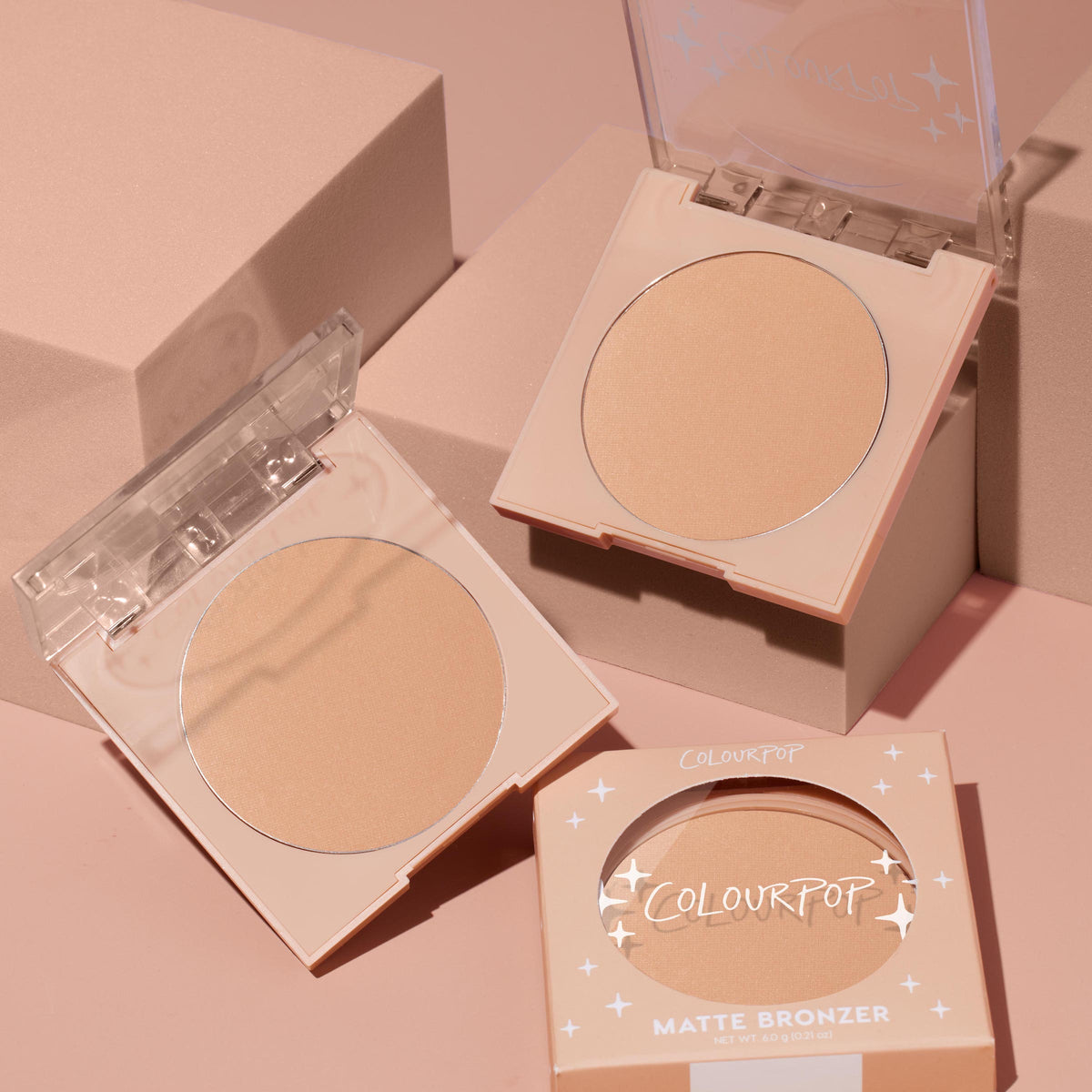 Avila Beach ColourPop Pressed Powder Bronzer compact opens to reveal a warm matte tan pan, with a second open compact and boxed matte bronzer nearby on soft pink blocks, showcasing clear flip-top lids and the 6.0 g pressed powder.