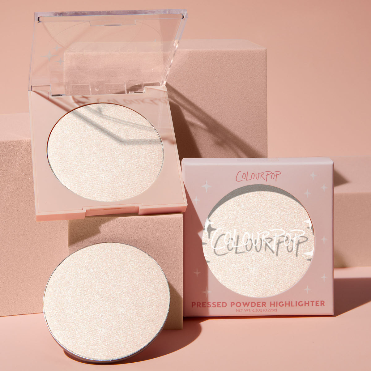 Cold One highlighter compact opens to reveal an icy pearl 0.23 oz pan, with a refill and star-print box beside it; ColourPop Pressed Powder Highlighter in a pale pink case styled on blush-pink blocks for a luminous glow.