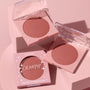 Just Cruisin’ ColourPop Pressed Powder Blush opens to reveal a warm rose shade in a pink compact with a clear lid, beside a second open pan and the retail box, styled on blush‑pink pedestals under soft studio lighting.