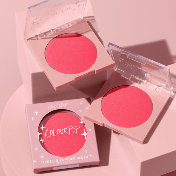 ColourPop Pressed Powder Blush Staycation, a bright coral-pink matte cheek color, shown in nude-pink square compacts with clear lids and one boxed, arranged on blush-toned blocks under studio lighting.