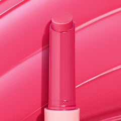 Tea Sesh gloss balm stick stands upright with the bright pink bullet extended and gleaming, set against sweeping hot-pink swatches; ColourPop So Juicy Plumping Gloss Balm with Peptides in a translucent pink tube with a light pink base.