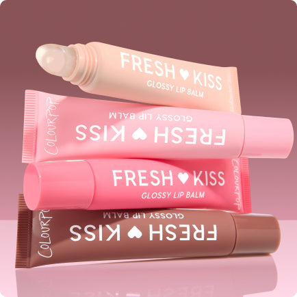 Colourpop Fresh Kiss Glossy Lip Balm tubes stacked diagonally, one open with a clear applicator, in peach, light pink, hot pink, and cocoa shades on a reflective pink surface against a mauve backdrop, highlighting hydrating lip care.