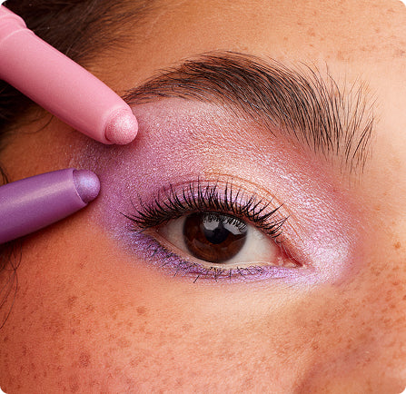 Two pastel Colourpop eyeshadow sticks in pink and purple glide onto a brown eye, creating a shimmery lavender‑pink look across the lid and lower lash line; close-up highlights creamy stick formula and luminous finish.