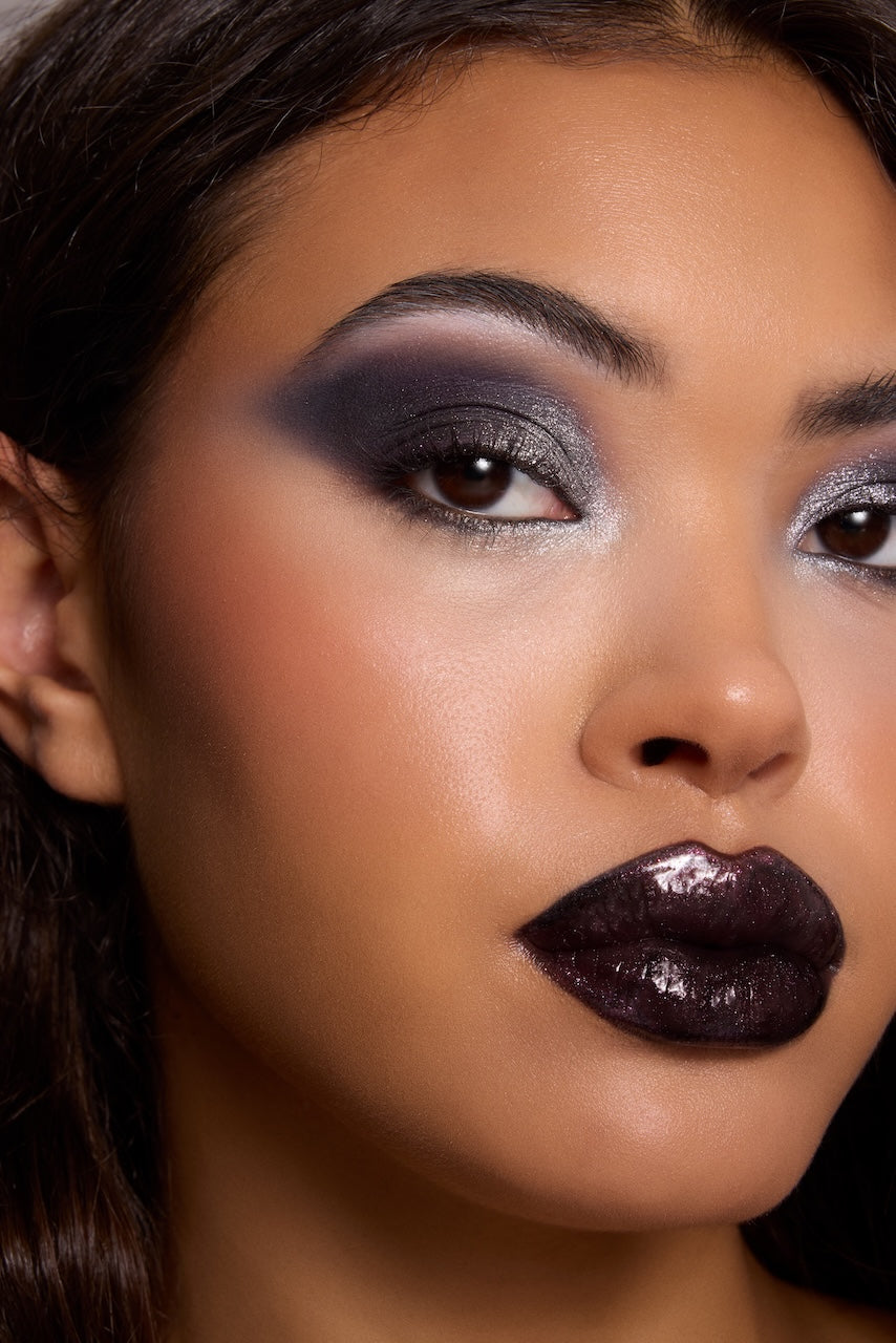 Bold smoky eye and glossy deep plum lips showcase ColourPop makeup, featuring silver‑gray shimmer eyeshadow blended to a wing and high‑shine black cherry lip gloss on a model in a studio close‑up beauty shot.