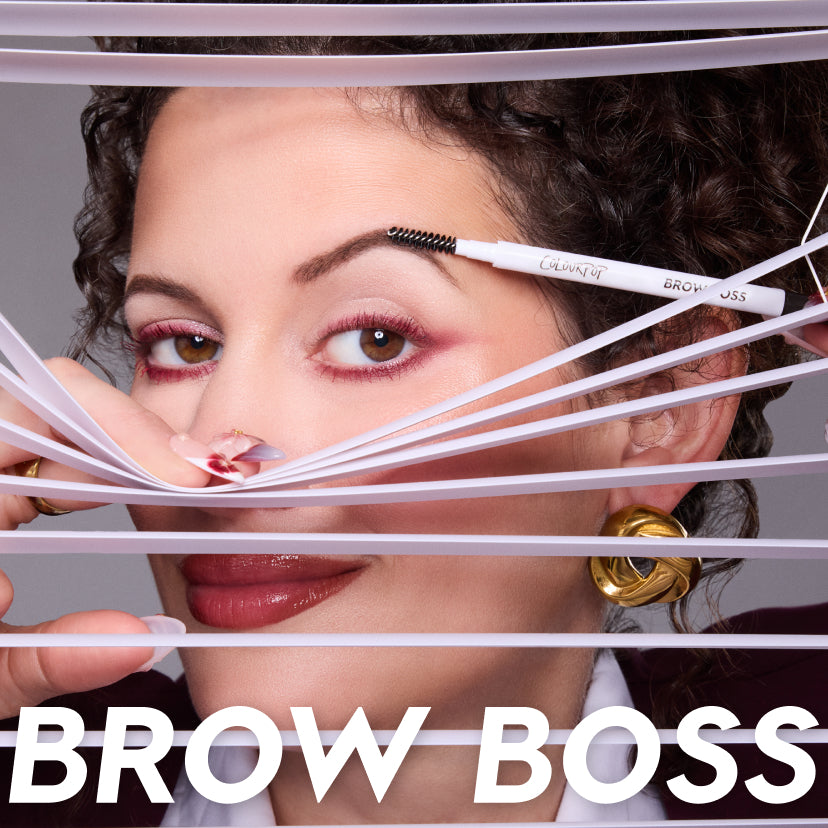 Woman grooming her eyebrow with the ColourPop Brow Boss pencil, peeking through white blinds, showcasing defined brows, bold pink eye makeup, glossy lips, and gold hoop earrings in a close-up beauty product shot