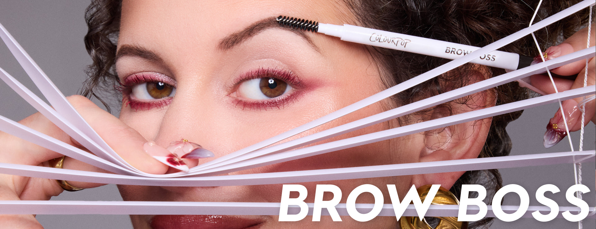 ColourPop Brow Boss eyebrow pencil brushing a woman’s perfectly groomed brows as she peers through white blinds, showcasing defined arches, pink eye makeup, and glossy nails in a close-up beauty product banner for US shoppers.