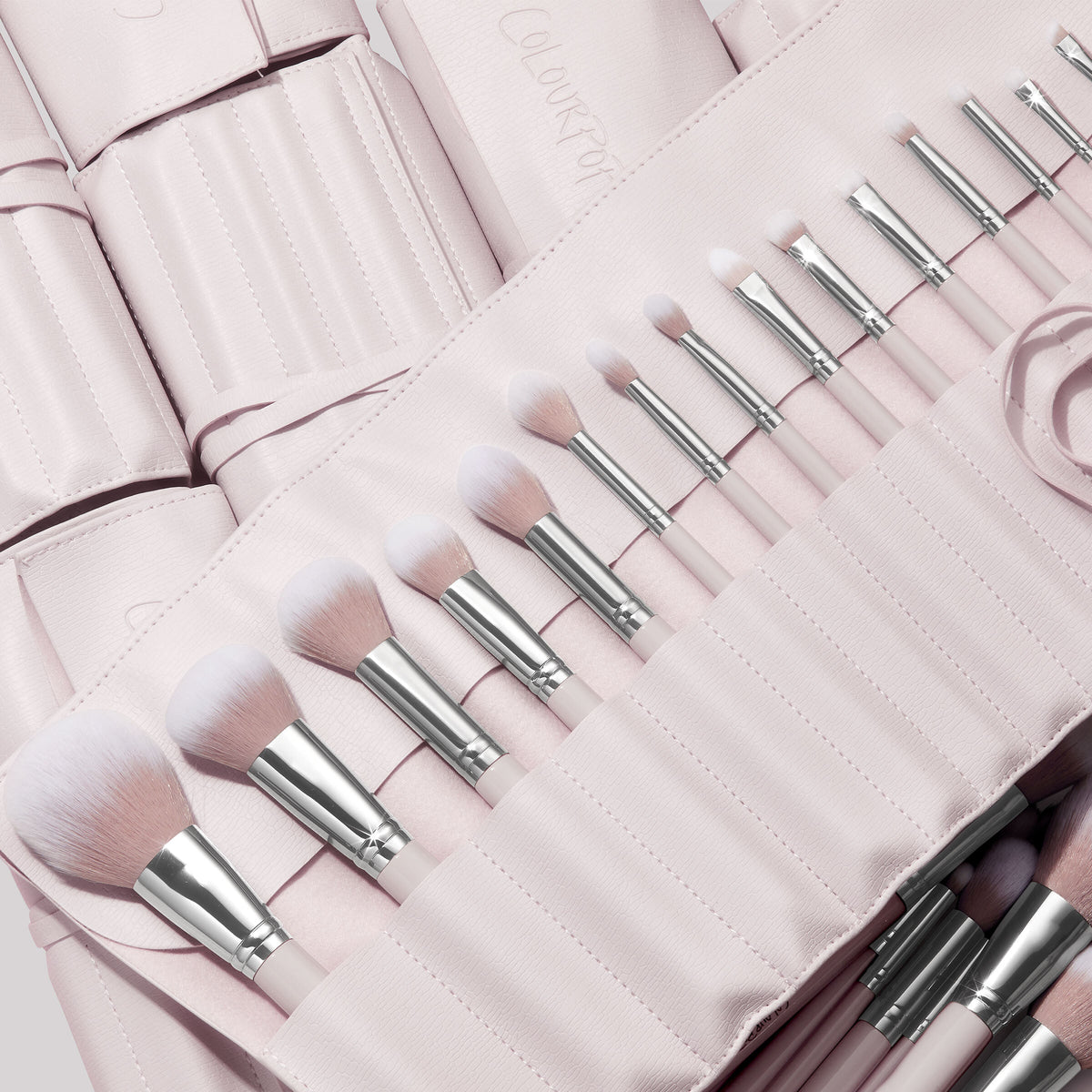 Stone Cold, a ColourPop Makeup Brush Kit, fanned in a blush-pink roll as face and eye brushes slot into stitched pockets, soft white-to-pink bristles and silver ferrules gleaming, with additional matching brush rolls layered behind.