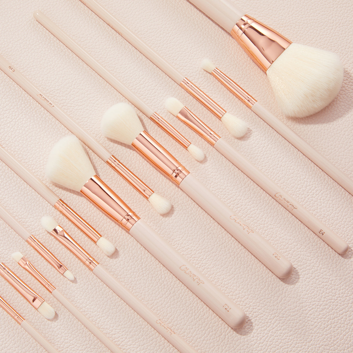 ColourPop Makeup Brush Kit lined up diagonally, the Ultimate Brush Roll features fluffy face and eye brushes with white bristles, rose-gold ferrules, and pale pink handles resting on a blush textured backdrop.