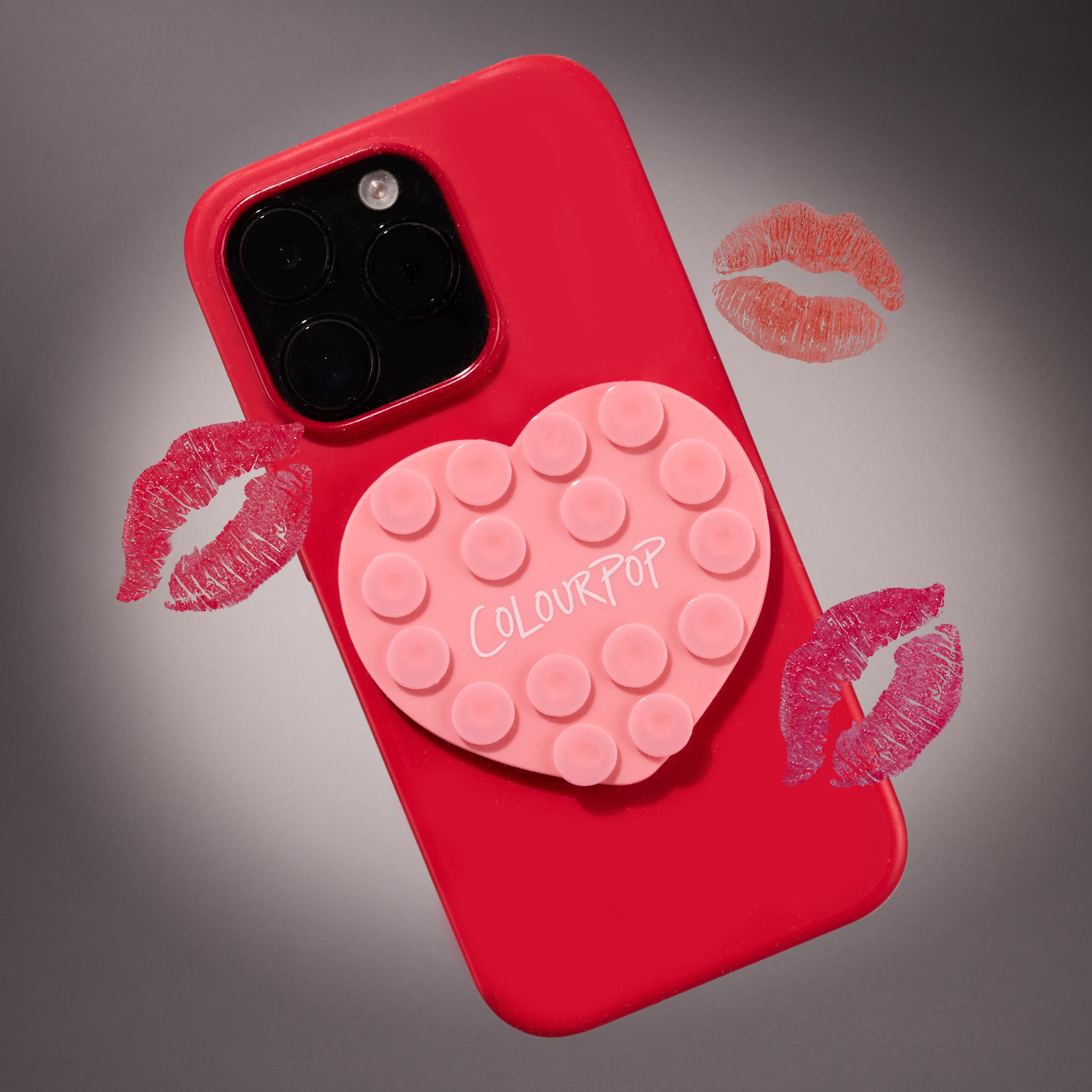 Phone Grippy heart-shaped pink suction grip branded ColourPop securing the back of a red smartphone case, surrounded by lipstick kiss marks on a gray gradient background; ColourPop GWP gift with purchase accessory.