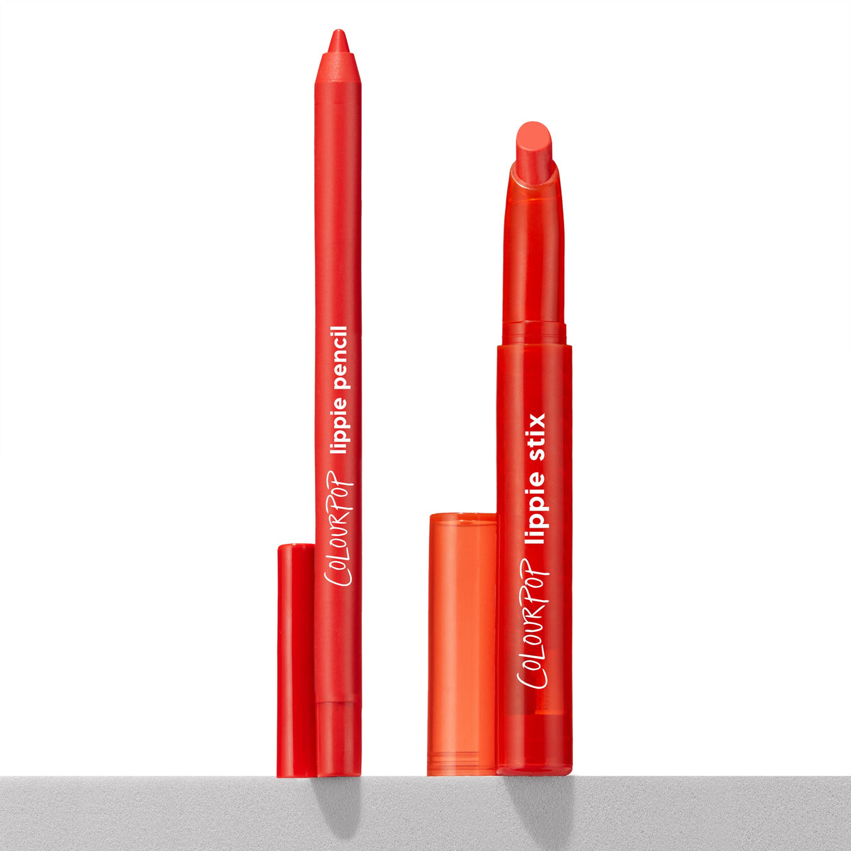 ColourPop Lippie Stix + Pencil Set Sealed with a Kiss standing uncapped, orange-red twist-up lippie stix and matching slim lip pencil showing tips, caps beside them on a white backdrop and gray ledge, for lining and filling lips.