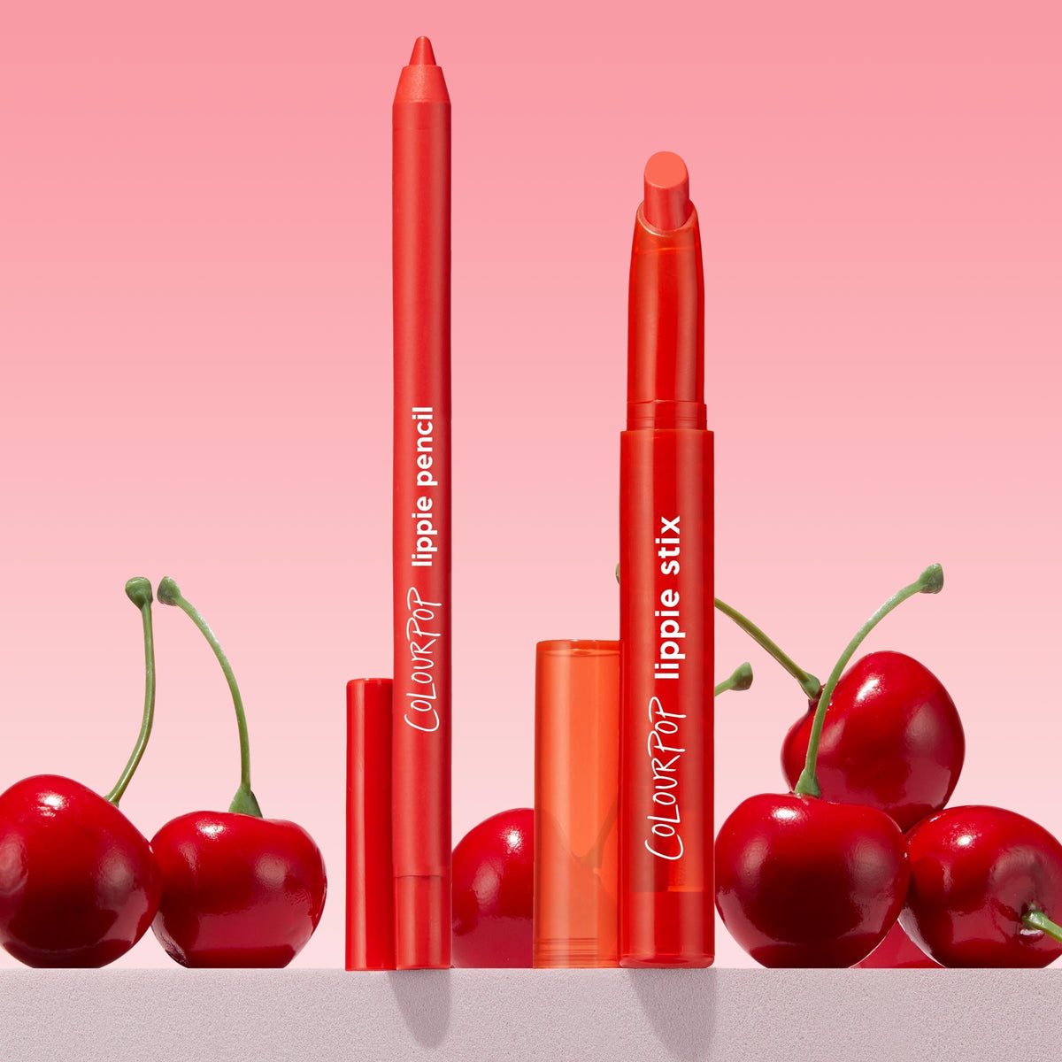 Sealed with a Kiss ColourPop Lippie Stix + Pencil Set stands uncapped, showing a creamy cherry-red Lippie Stix and matching lip pencil, posed amid fresh cherries on a light surface against a pink gradient background.