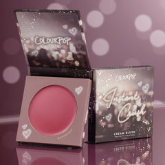 ColourPop Cream Blush in Obviously opens to show a rosy pink pan, with the Instant Crush box propped beside it; heart-and-bokeh graphics decorate the packaging on a reflective mauve backdrop.