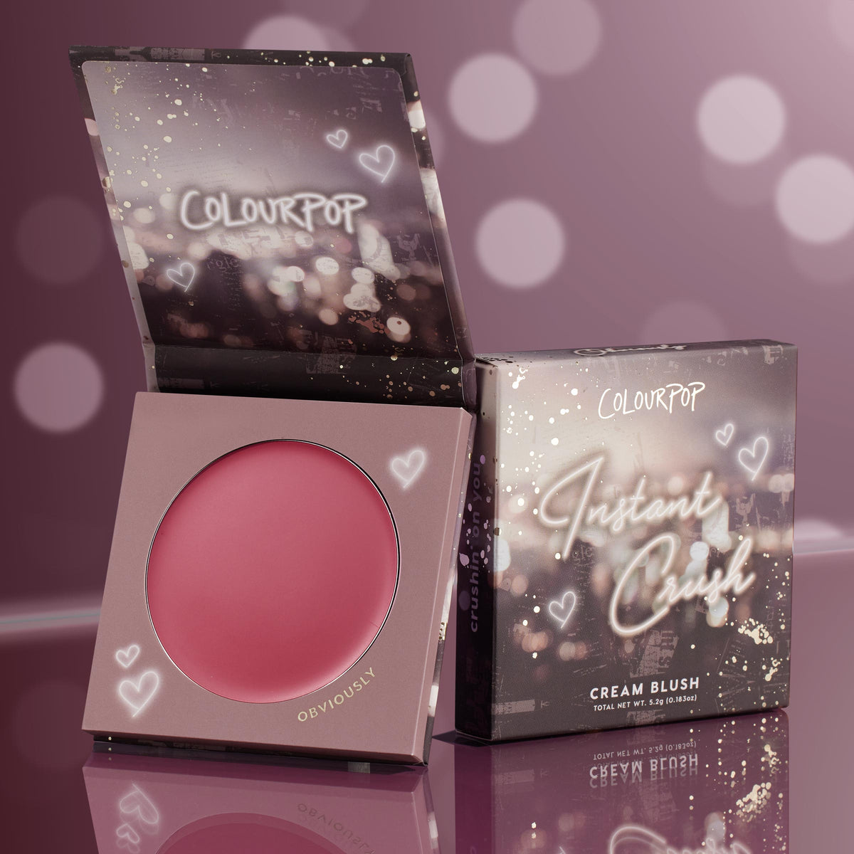 ColourPop Cream Blush in Obviously opens to show a rosy pink pan, with the Instant Crush box propped beside it; heart-and-bokeh graphics decorate the packaging on a reflective mauve backdrop.