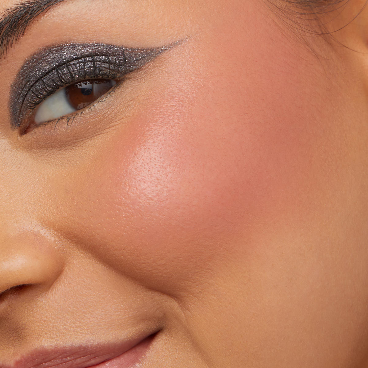 Cheek wearing ColourPop Cream Blush in shade Obviously, showing a soft peach flush with a dewy finish as the model smiles; smoky winged eye makeup visible in a tight close-up for product application detail.