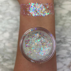 Disco Lady glitter gel swatched across a forearm, displaying chunky iridescent rainbow sparkle; an open pot of translucent gel packed with holographic flakes rests on the arm over a marble surface, showcasing payoff and texture; ColourPop Glitter Gel.