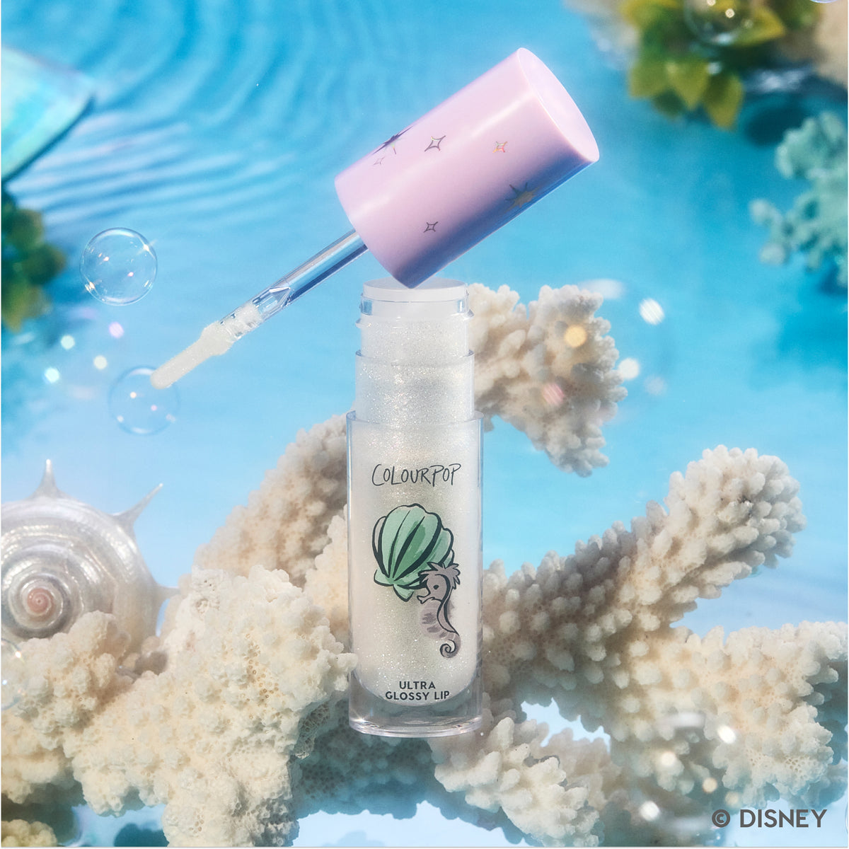 ColourPop Bright Ariel Ultra Glossy Lip tube with wand lifted, showing shimmering clear gloss in an underwater-inspired setting with coral, bubbles, and seashells, evoking a Disney Princess mermaid theme for US beauty fans.