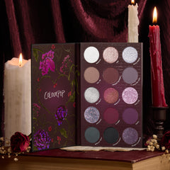 A Shadow of Secrets ColourPop Shadow Palette opens to 15 pans of cool-toned mauves, burgundies, and silver shimmers in a floral case, staged on a vintage book with lit candles and dried roses for a moody, velvet-draped setup.