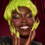 ColourPop How the Grinch Stole Christmas shadow palette showcasing shimmery olive-green lids on a smiling model, eyes closed, with neon green wig and glossy brown lips, set against a deep red velvet backdrop.